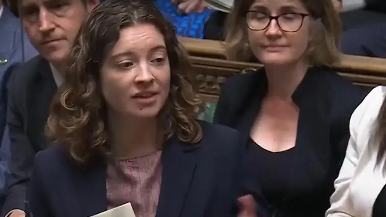 The MP, who has dark brown curly hair, is speaking Parliament. She has a dark jacket on over a pink top. A male and female MP can be seen behind her.