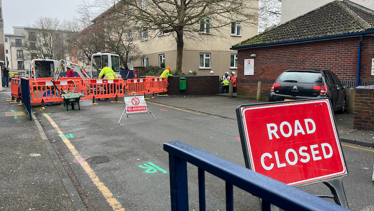 A red ROAD CLOSED sign in front of a fenced off part of a road surrounded by residential buildings with people in high vis gear working.