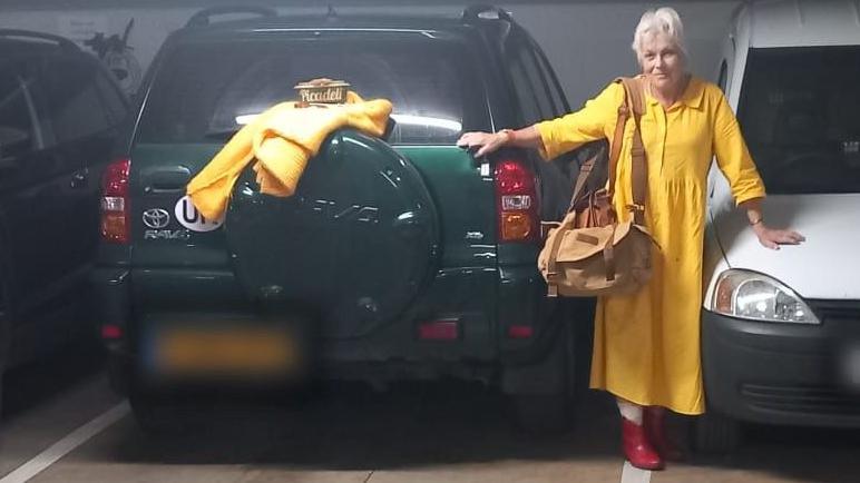 The image shows an indoor parking garage a green SUV with a yellow item draped over its spare tyre. A person wearing a yellow coat and red shoes is standing between the green SUV and a white van, holding onto both vehicles. 