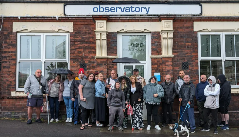 A group of 19 people stand outside a brick building which has a sign that says Observatory. Some of the crowd are holding umbrellas and one of them has a dog