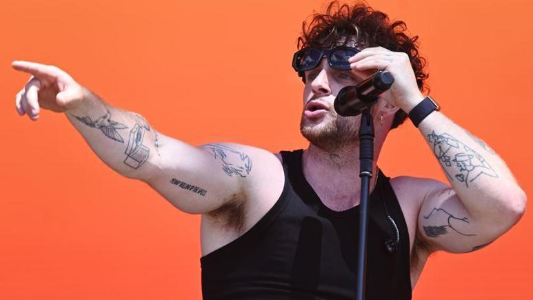 Tom Grennan, with dark hair and tattoos, wearing a vest top on stage sings into a microphone 