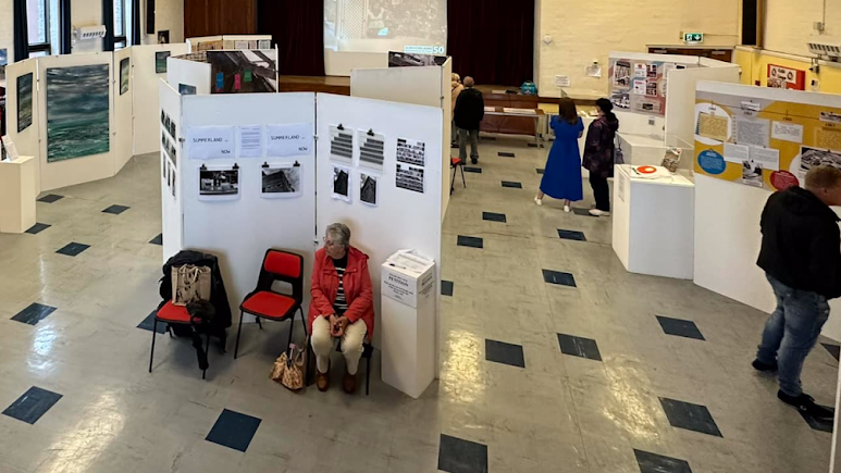 The exhibition seen from above. There are people looking at white boards featuring pictures and information positioned on a black and white tiled floor.