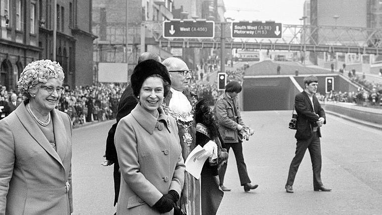 A smiling Queen Elizabeth is pictured with other dignitaries and photographers at the entrance to tunnels named Queensway at Great Charles Street. She is wearing a dark fur hat, collared coat and black gloves. She is standing next to a lord mayor wearing his ceremonial robes. A smiling lady also stands next to her in a jacket, pearls and striking hat.
