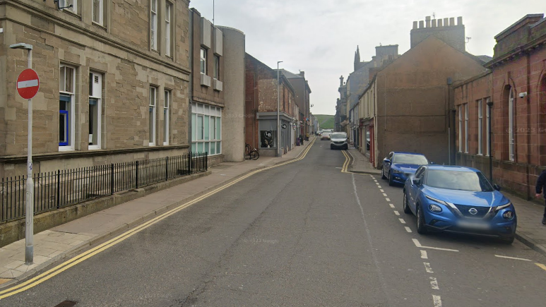 A general view of Commerce Street in Arbroath. There is a stop sign and two parked blue cars. There are several buildings on either side of the road. A van is parked on the right hand side in the distance.