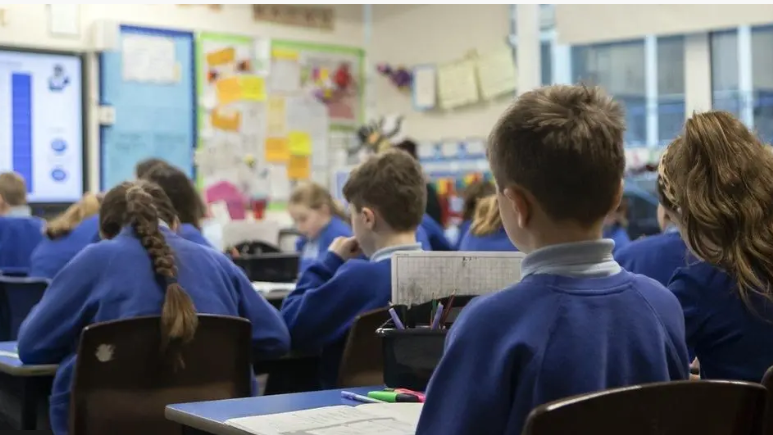 Clann-sgoile ann an seòmar-teagaisg. Tha lèintean gorma orra. 