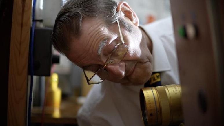 Pat Church wearing a smart shirt and tie while inspecting the lens on a gold projector