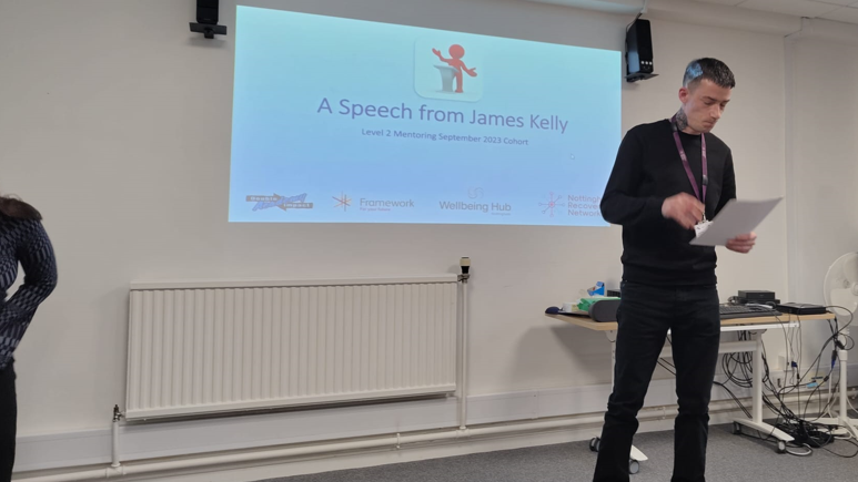 Mr Kelly is wearing a black top and dark jeans and is stood up addressing people in a room with a screen behind him.