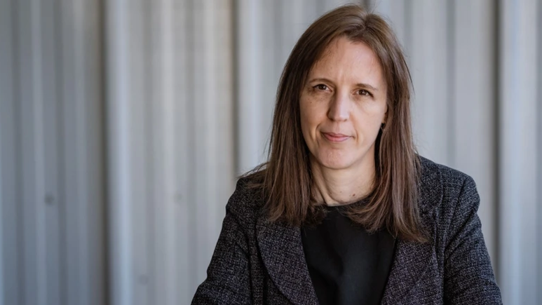 Catherine Braun in a grey suit sat in front of a grey background looking in to the camera