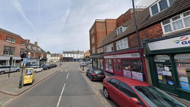 Google street image of Grantham town centre. Several shops can be seen on either side of the road and several parked cars with a market place and buildings in the distance.