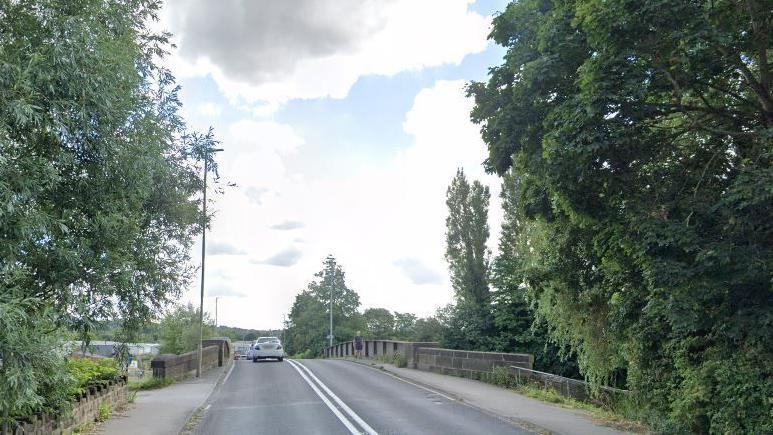 A road which leads onto a bridge with a car on it and trees on either side 