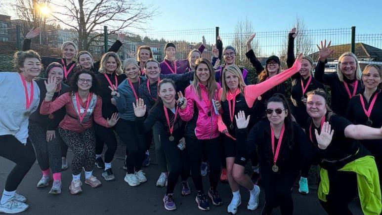 Members of the club are gathered together for a group photograph. They are all wearing running gear and have pink lanyards and medals. There is a fence in the background with winter trees and a blue sky.
