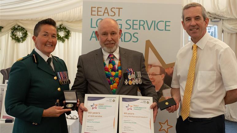 Clive Parnham standing in the centre of a group of three people. He is looking directly at the camera and smiling. He is wearing a grey suit with a colourful waistcoat and matching tie. He is also holding up two certificates which he has been awarded.