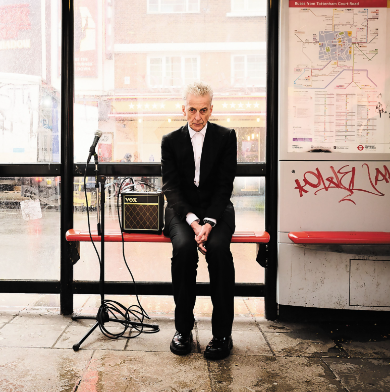 The cover of the album Sweet Illusions shows Peter Capaldi in a black suit and white shirt sitting on a bus shelter seat in London with a microphone stand and amp beside him