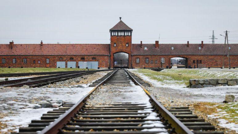 Train tracks leading to a red brick building extending across the frame. Alongside the tracks are patches of grass and gravel covered in snow