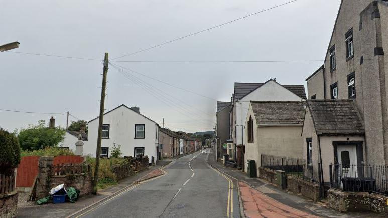 The A5086 Main Street in Cleator: terraced houses with slate roofs line the road on either side; there is a white car is in the distance.