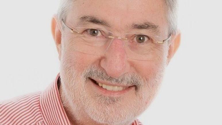 Ray McMahon has a a red and white striped shirt. He wears small spectacles, has short grey hair and white stubble.