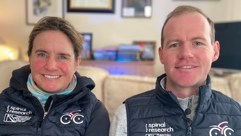 Piggy and Tom March sitting side by side on a cream sofa, both wearing dark blue gilets with Spinal Research and British Eventing Support Trust logos on one side of the chest, and a cycling logo on the other. A table lamp, sideboard, framed pictures on the wall and a TV can be seen behind them. 