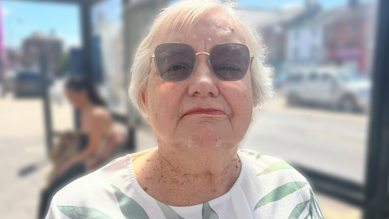 Diane Quadling, a woman with white, short hair, is looking into the camera. She is wearing tinted glasses, and a white top with a green, leafy pattern on it.