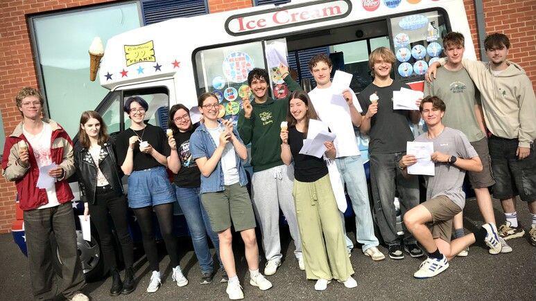 A large group of students all stand in front of an ice cream van. They are all grinning widely at the camera while holding their results papers in one hand and a Mr Whippy ice cream in the other.