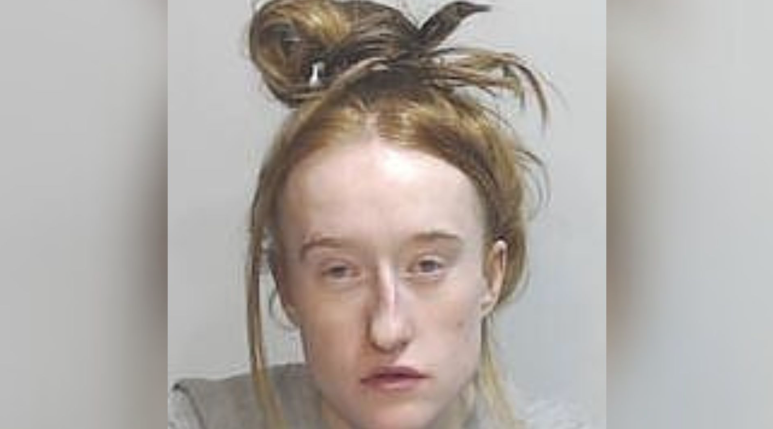 a police mugshot of a woman with fair hair tied up on top of her head with strands coming down at the side of her face