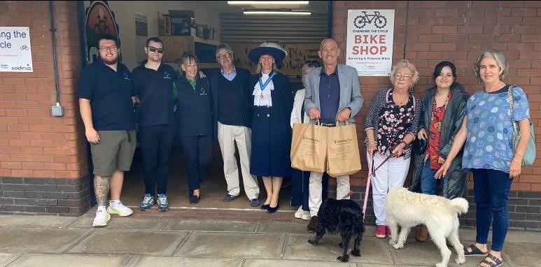 There are 10 people pictured in front of the shop including the High Sheriff of Northumberland, Lucia Bridgeman, who is wearing a traditional hat with a feather. There are also two dogs. On the wall are signs saying Bike shop and Changing the Cycle.