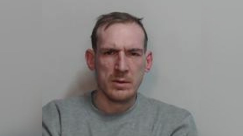 a police mugshot of a man with short brown hair and wearing a grey sweatshirt