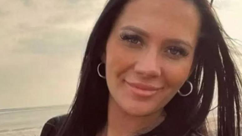 Kiena Dawes with long black hair and silver hoop earrings on a beach smiling.
