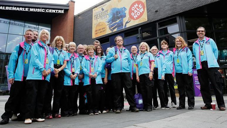 A group of about 30 volunteers, of all ages and both male and female, are standing outside Albemarle Music Centre in Hull city centre. They are all wearing sky blue coats that became synonymous with the 2017 City of Culture volunteers.