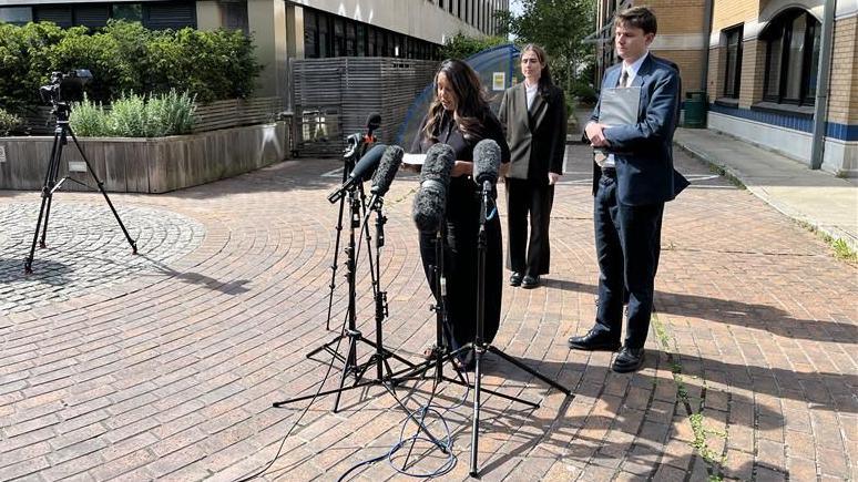 A woman dressed in all black reading from a sheet of paper. She is standing in front of a group of microphones.