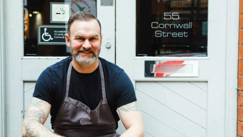 A man with short, dark hair and a beard, is wearing a black T-shirt and a grey apron, sits on the steps outside a building. One of the glass doors is labelled with "55 Cornwall Street".