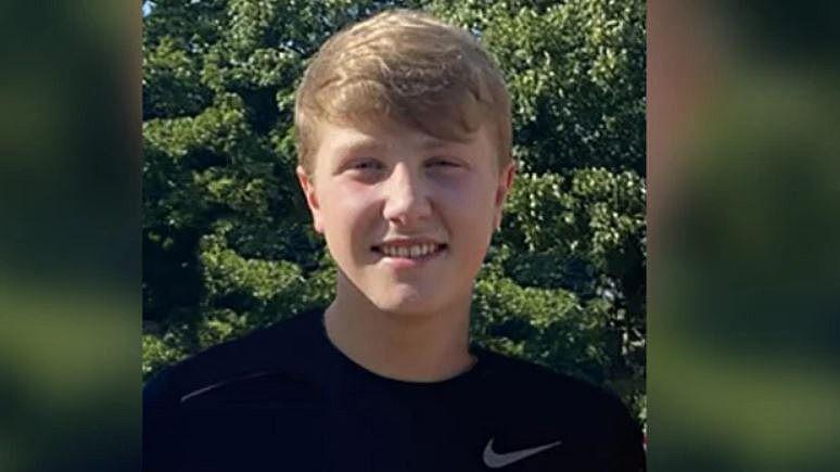 Matthew Daulby with blond hair wearing a black t-shirt is standing in front of trees on a sunny day. He is smiling.