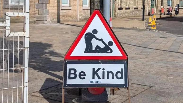 A red triangle road hazard sign, with an outline cartoon character picking another up from the floor, with the wording "Be Kind" on it.