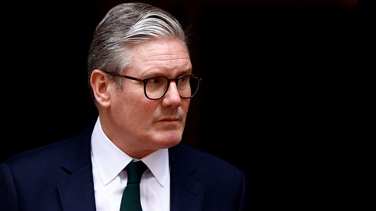 A head-and-shoulders picture of Keir Starmer. He is wearing a dark blue suite, a white shirt and a dark tie. He has a serious expression on his face. He is pictured against a dark background. 