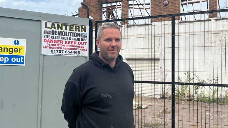 Business owner Craig Ross, who has grey hair and is wearing a dark hooded top, stands in front of a fence that has been erected around the wreckage of the industrial estate.