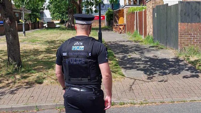Police officer wearing black shirt and trousers and black cap with white and black braiding. The word POLICE is on the back of the shirt. He has his back to the camera and is walking on a block-paved path towards a group of two-storey terraced houses. There is a grassed area immediately in front of him and a tree, the branches of which are hanging above the pavement.