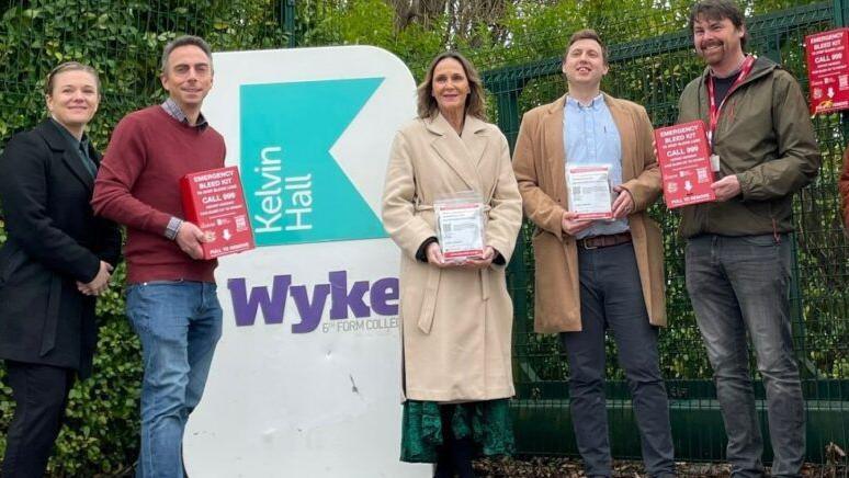 A group of five men and women pose for a photo with the new bleed kits next to a sign outside Kelvin Hall School, there is a green fence and shrubbery in the background.