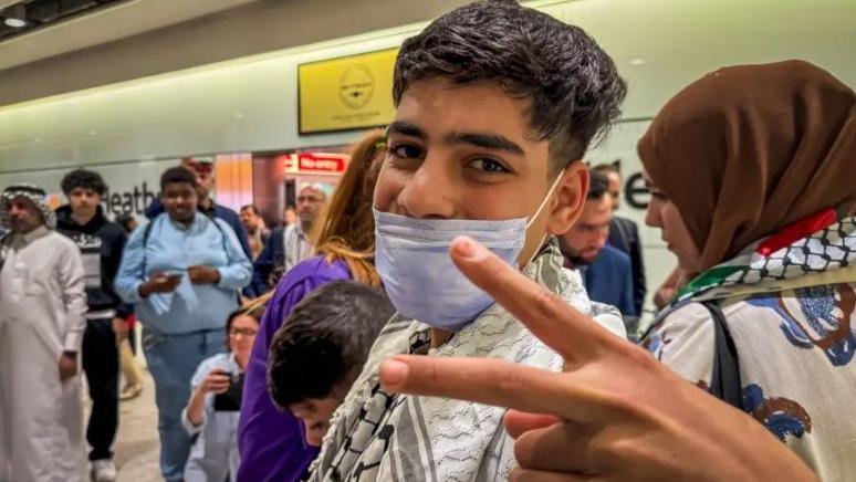 Majd is a teenage boy, wearing a facemask over his lower face. He is in a busy Heathrow arrivals hall and making a victory sign towards the camera