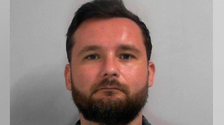 A police custody image of a man with short dark-brown hair, brown eyes and a trimmed brown beard. He has a serious expression as he looks into the camera against a plain light-grey backdrop. He is wearing a blue and white plaid shirt over a white t-shirt.