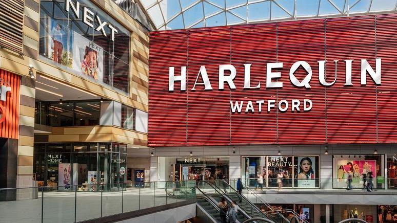 Red internal wall of the Atria centre, with lettering saying "Harlequin Watford". The Q in Harlequin has a diamond-shaped "hole" in the middle, reflecting the traditional Harlequin checked pattern. There is a next store below the sign, featuring pictures of models wearing Next clothing. There is an escalator in the foreground and another shop to the left of the picture.