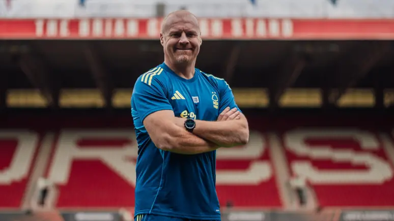 Sean Dyche standing on the Nottingham Forest pitch with his arms folded. He is wearing a blue, short-sleeved football shirt. The empty stand, which is out of focus, can be seen g=behind him
