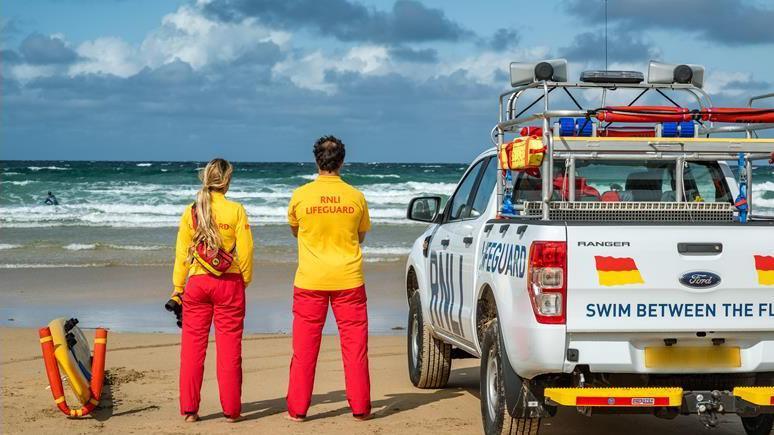 Two lifeguards in yellow T-shirts and bright red trousers stand together, looking out to sea. 