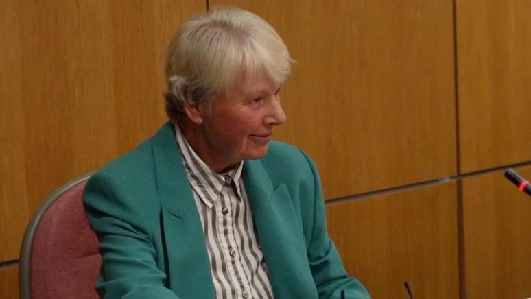Rosemary Thew sits at a desk inside the council chamber. She is wearing a green jacket, striped black and white shirt and has white hair.