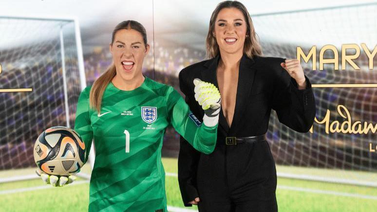 Waxwork Mary Earps in her green England goalkeeper kit with the real-life Mary Earps. She is wearing a black jump suit and is mimicking her waxwork with a raised fist to the camera. 