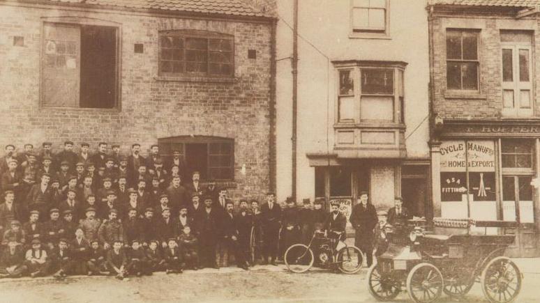 A black and white archive photo of men standing outside a one-storey brick building. One of the men at the front is holding a bicycle. There is a shop on the left that has a sign saying "F Hopper Cycle Manufacturer".