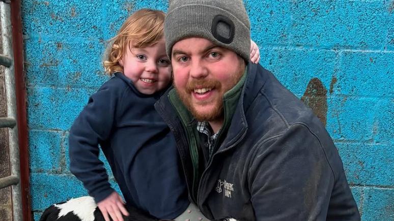 Andrew Little and daughter Molly in navy boiler suits, smiling holding a black and white calf, pictured against a bright blue wall in a farm shed