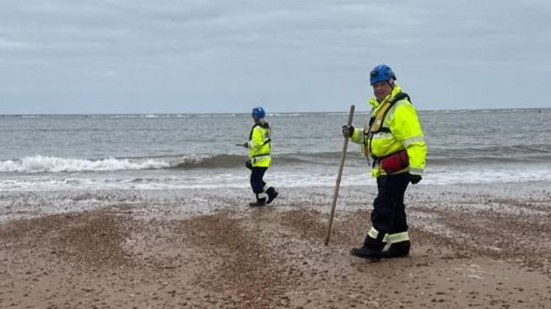 Two HM Coastguard employees walking on a beach, both wearing high vis and blue helmets, looking for nurdles - small plastic pellets. The one closest to the camera is holding a long stick.