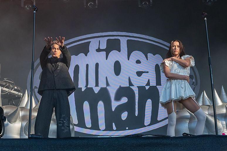 A man in a baggy black suit holds his arms towards the crowd. He is stood next to a woman in a white dress and white tights. The words confidence man are spelled out on a screen behind the pair