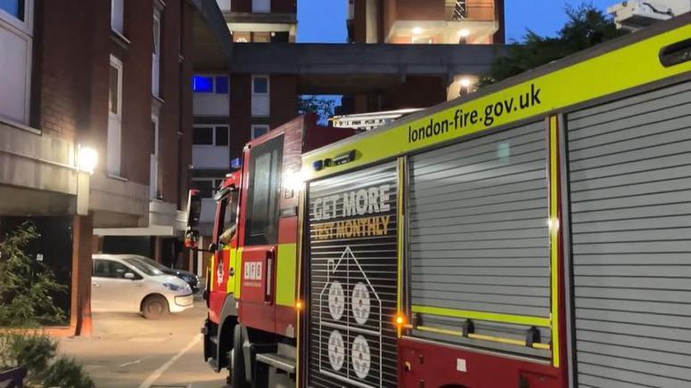 A red and yellow fire engine sits parks in the carpark of the block of flats. The block is made of brick and concrete and features white UPVC windows