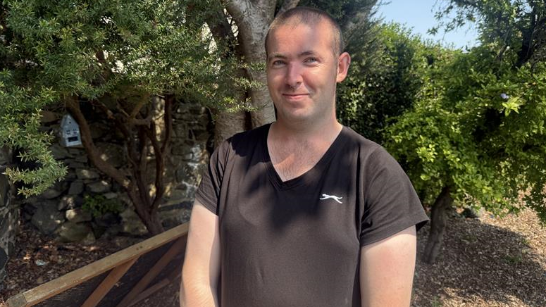 A smiling man in a black t-shirt standing in front of a small group of trees.