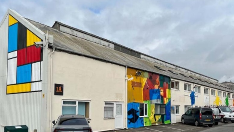 A long warehouse type building with colourful murals on the walls and some cars parked at the front